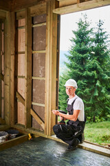 Carpenter constructing wooden framed house. Man worker working with screwdriver, wearing work overalls and helmet. Concept of modern eco-friendly construction.