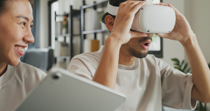 Young Asian couple sit on couch hold digital tablet and wear virtual reality glasses are watching and show imagine spend time together have fun at home on weekend. Lifestyle tech innovation concept.