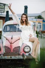 Fototapeta na wymiar A beautiful young girl in a white dress posing near red retro car. Summer photo. Spring fashion model concept. Vintage and retro style. Luxury travel.