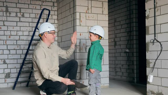 A little boy high-fives his dad at a construction site. Dad and son in white helmets at the construction site are happy to get a new apartment. High quality 4k footage