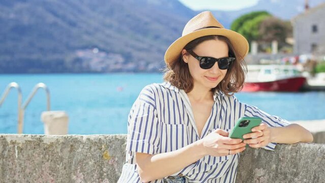 A young Caucasian woman in a hat and sunglasses using her phone while sitting by the sea on a sunny day.