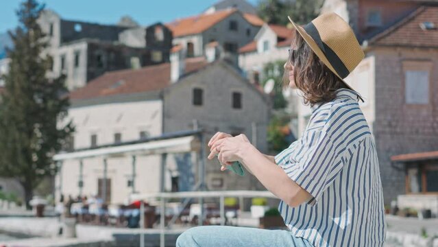 A young Caucasian woman in a hat taking a picture with her phone while sitting against the background of an old European seaside town