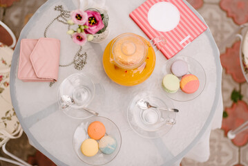 Summer cafe table. On the table are glass plates, cups, a kettle with juice, tea utensils. There are macaroons of different colors in the plates..On the table is a woman's handbag and a notebook