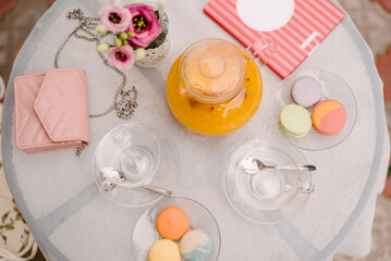 Summer cafe table. On the table are glass plates, cups, a kettle with juice, tea utensils. There are macaroons of different colors in the plates..On the table is a woman's handbag and a notebook