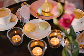 Romantic evening in a cafe..Three candles are burning, two macaroons and desserts are on the plate, and cutlery is next to it on a white napkin. Two cups of coffee on the table