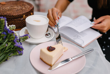 On the table of the summer cafe lies a women's handbag woven from a wicker and an open book that the girl is reading. Dessert with strawberries on a pink plate. Breaks dessert with a fork