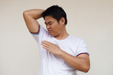 Asian man checks his body odor during he feels hot, raises right arm up, smells his armpit,...