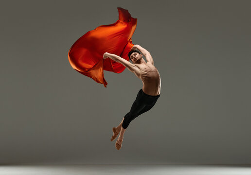 Young artistic man, ballet dancer with muscular body dancing, making performance with red silk fabric on grey studio background. Concept of art, classical dance, inspiration, creativity, choreography