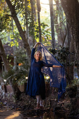 A beautiful fifty-year-old woman with light hair, dressed in a blue dress, is looking into the frame. The scene around her consists of nature during sunset. This is an anti-aging concept 