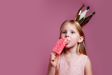 Cute little girl in a fancy dress and a crown eating ice-cream on pink background
