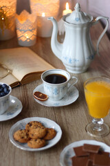 Cup of tea or coffee, plate of cookies, cup of blueberries, plate of chocolate, glass of juice, book, reading glasses, teapot, flowers and lit candles on the table. Brunch or afternoon tea concept.