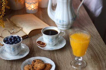 Cup of tea or coffee, plate of cookies, cup of blueberries, plate of chocolate, glass of juice, book, reading glasses, teapot, flowers and lit candles on the table. Brunch or afternoon tea concept.