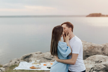 Happy couple drinking white wine at picnic. Young couple enjoying picnic time on the sunset. A boyfriend and a girl on a romantic date kissing. Copy space.