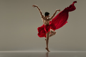 Young, handsome, muscular man dancing with red silk fabric against grey studio background. Modern ballet performance. Concept of art, classical dance, inspiration, creativity, fashion, choreography