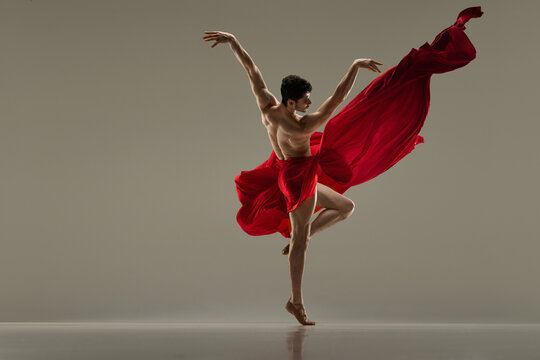 Young, handsome, muscular man dancing with red silk fabric against grey studio background. Modern ballet performance. Concept of art, classical dance, inspiration, creativity, fashion, choreography