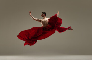 Artistic, talented, young man, shirtless ballet dancer dancing with red silk fabric against grey studio background. Concept of art, classical dance, inspiration, creativity, beauty, choreography