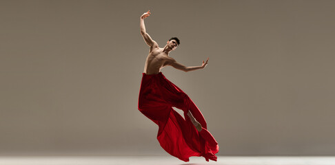 Freedom. Talented, artistic young guy, professional ballet dancer performing shirtless with red fabric around body on grey studio background. Concept of art, classical dance, inspiration, choreography - obrazy, fototapety, plakaty
