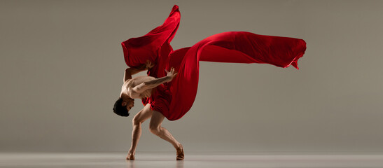Talented performance. Handsome man, ballet dancer dancing with red silk fabric against grey studio...