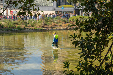 Sanitation worker for pest control dressed in helmet, goggles, mask with filters, protective gloves and boots pouring disinfectant for mosquitoes with a bucket in the pond of the Sagrada Familia