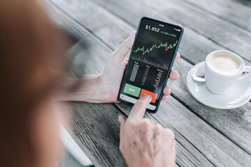 Over shoulder view of woman using mobile phone app to sell crypto currencies. Digital senior people and stock market concept