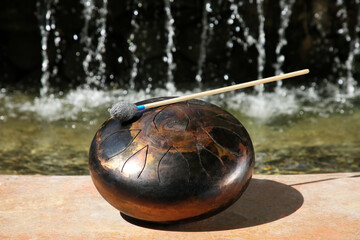 Steel tongue drum with mallet near waterfall outdoors on sunny day. Percussion musical instrument