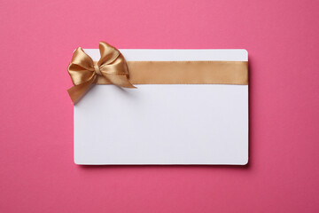 Blank gift card with golden bow on pink background, top view. Space for text - obrazy, fototapety, plakaty