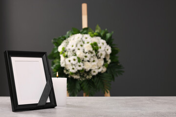 Photo frame with black ribbon, burning candle on light table and wreath of flowers near grey wall...