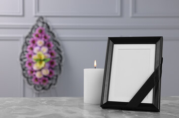Photo frame with black ribbon, burning candle on light grey table and wreath of plastic flowers near wall indoors, space for text. Funeral attributes