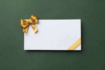 Blank gift card with golden bow on dark green background, top view