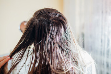 Woman checking dirty oily and greasy hair looking in mirror at home. Bad hair care cosmetics concept.
