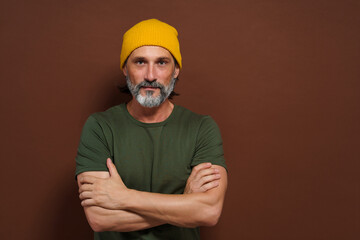 Mature, handsome man with silver beard wearing yellow hat against brown background. The image has copy space and can be used for product placement or advertising concepts.