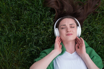 A teenage girl lies on the grass in white headphones, her eyes are closed, listening to music,...