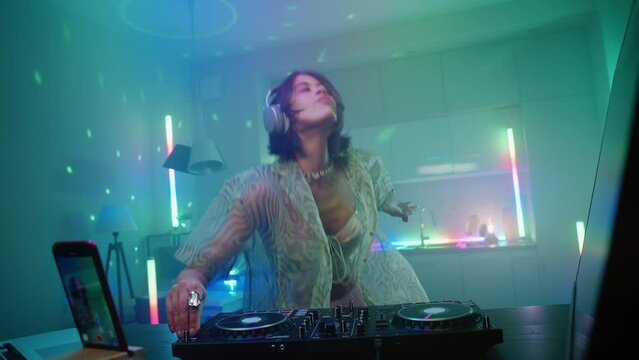 Beautiful young stylish woman dancing enjoying her live dj set in home interiors at night, excited female in headphones recording electronic music on turntable console. Colored neon lights glow
