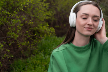 A young woman sits in the green spring grass and enjoys music in the park, listening in headphones with her eyes closed and a blissful smile, dressed in a green body shirt