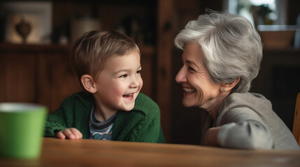 A grandmother and her young grandson sit side by side at a cozy wooden table, each with a playful and joyful expression on their faces.