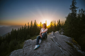 A girl is sitting on a boulder, the top of the rock. Sunset on a summer day. The tops of pine trees. The concept of active recreation, hiking and travel. Beautiful view of the mountains.