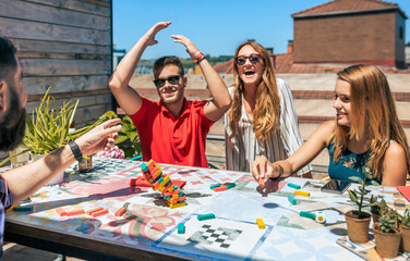 Group of happy friends laughing while a young man lose in jenga game in a rooftop party on summer