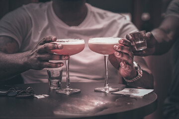 Hands Holding Two Cocktails in Buzzing Restaurant: Diverse Socializing, Refreshing Beverages, Vibrant Atmosphere, Nightlife, Enjoyment, Fun Dining Experience