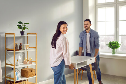 Happy family couple moving furniture at home. Joyful, positive young man and woman buy a new desk and put it in the living room in their modern apartment
