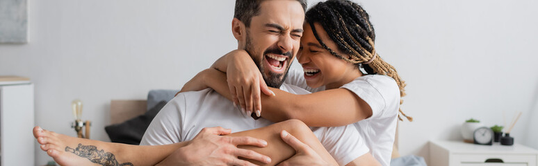 Fototapeta premium overjoyed interracial couple in white t-shirts embracing and laughing with closed eyes in bedroom at home, banner.