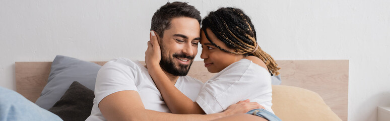 Obraz premium young african american woman with dreadlocks embracing joyful bearded boyfriend in bedroom at home, banner.