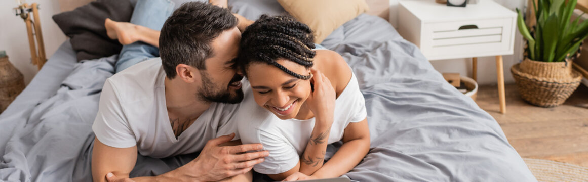 cheerful man telling secret to young african american woman while relaxing on bed at home, banner.