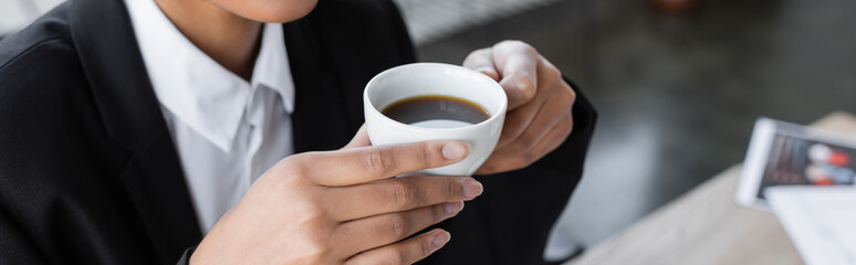 partial view of african american businesswoman holding cup of black coffee in office, banner.