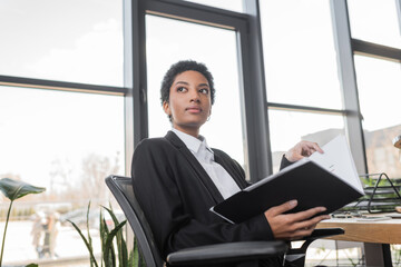 pensive african american businesswoman with folder sitting and looking away in office.