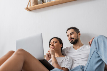 low angle view of smiling multiethnic couple watching movie on laptop while resting in bedroom at home.