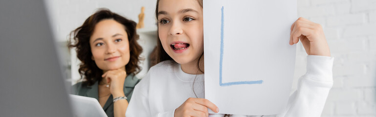 Child sticking out tongue and holding paper with letter during speech therapy online near mother at home, banner.