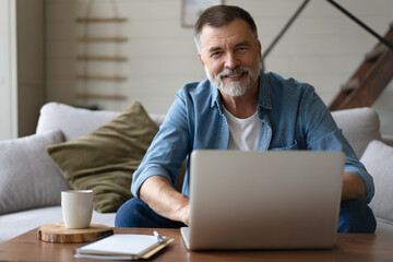 Senior man in casual clothing using laptop and smiling while sitting on the sofa, working from home.