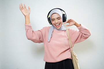 Smiling Asian Muslim female student in pink sweater with bag and listening to music excitedly isolated on white background. back to school concept
