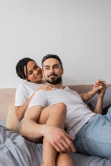 happy interracial couple in white t-shirts holding hands while lying on bed at home.
