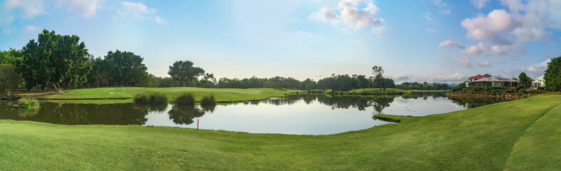 Wide panoramic view of premium lush golf course with beautiful pond in the foreground, surrounded by green grass and trees. 
